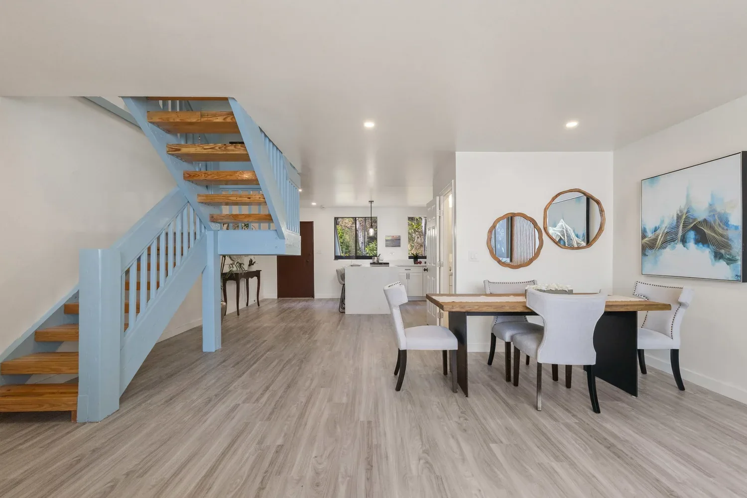 Open-concept living room and dining area with flooring, white walls, a staircase with light blue railing and wooden steps, and a kitchen in the background with a white marble island, white cabinetry, and black hardware. Decor includes wall art, mirro