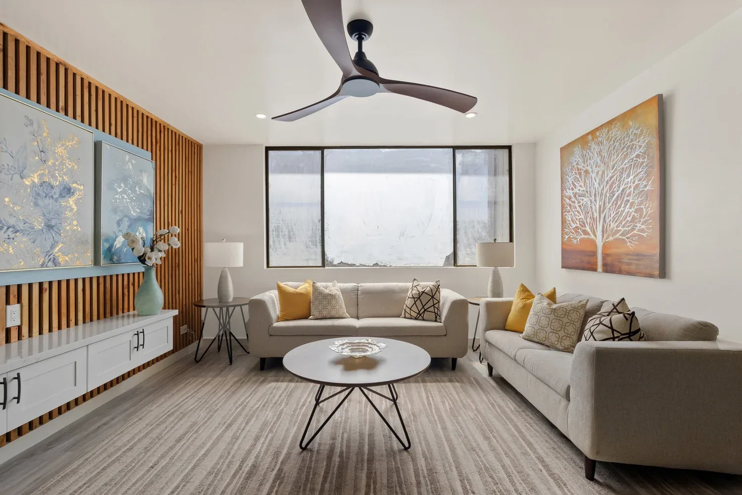 Modern living room with two light-colored sofas, a round coffee table, a wooden slatted accent wall, abstract paintings, floor lamps, and a large window.