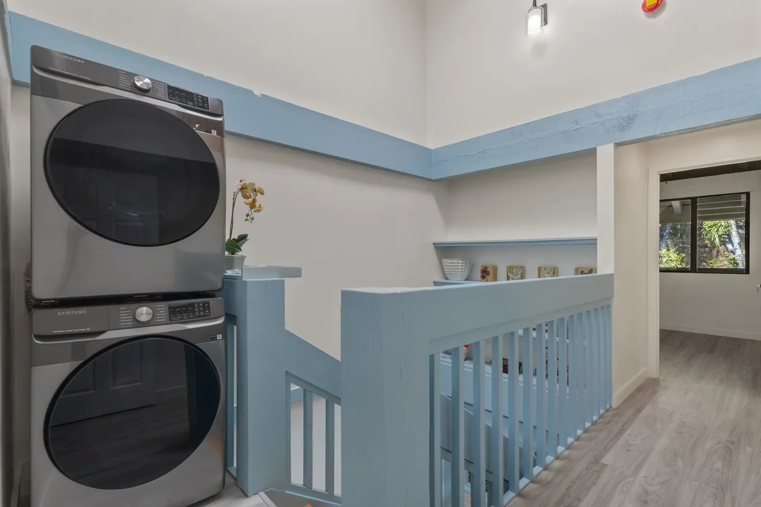 Laundry area with a stacked Samsung washer and dryer, a small potted plant, and a blue railing next to a staircase, in a bright room with a window showing greenery outside.