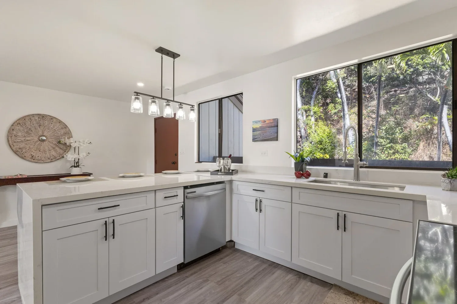 Modern white kitchen with large window overlooking trees, island with placemats, and hanging pendant light fixture.