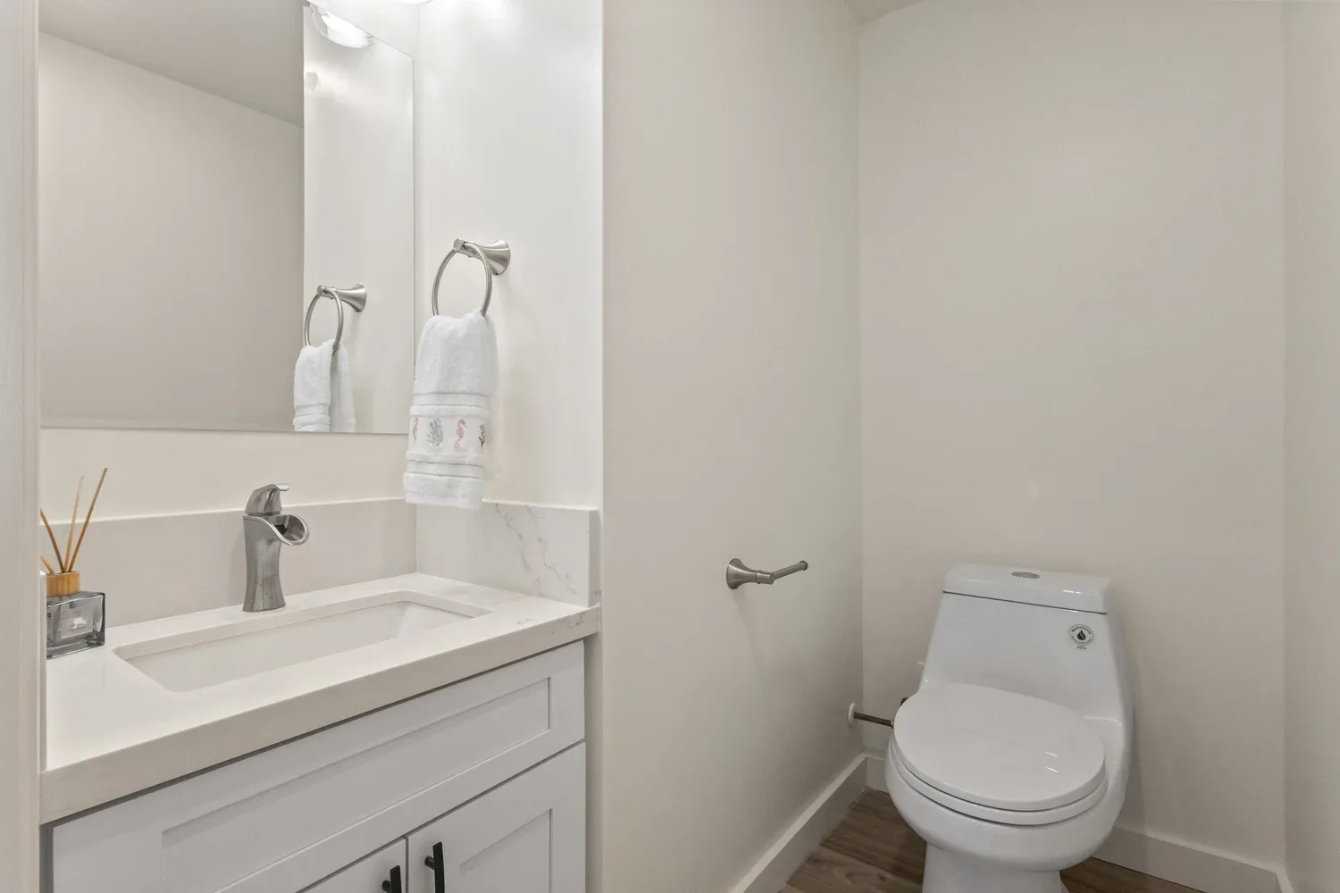 A small bathroom with a white vanity sink, a large mirror, a wall-mounted hand towel ring with a white towel, a reed diffuser, and a white toilet next to a towel holder on the wall.