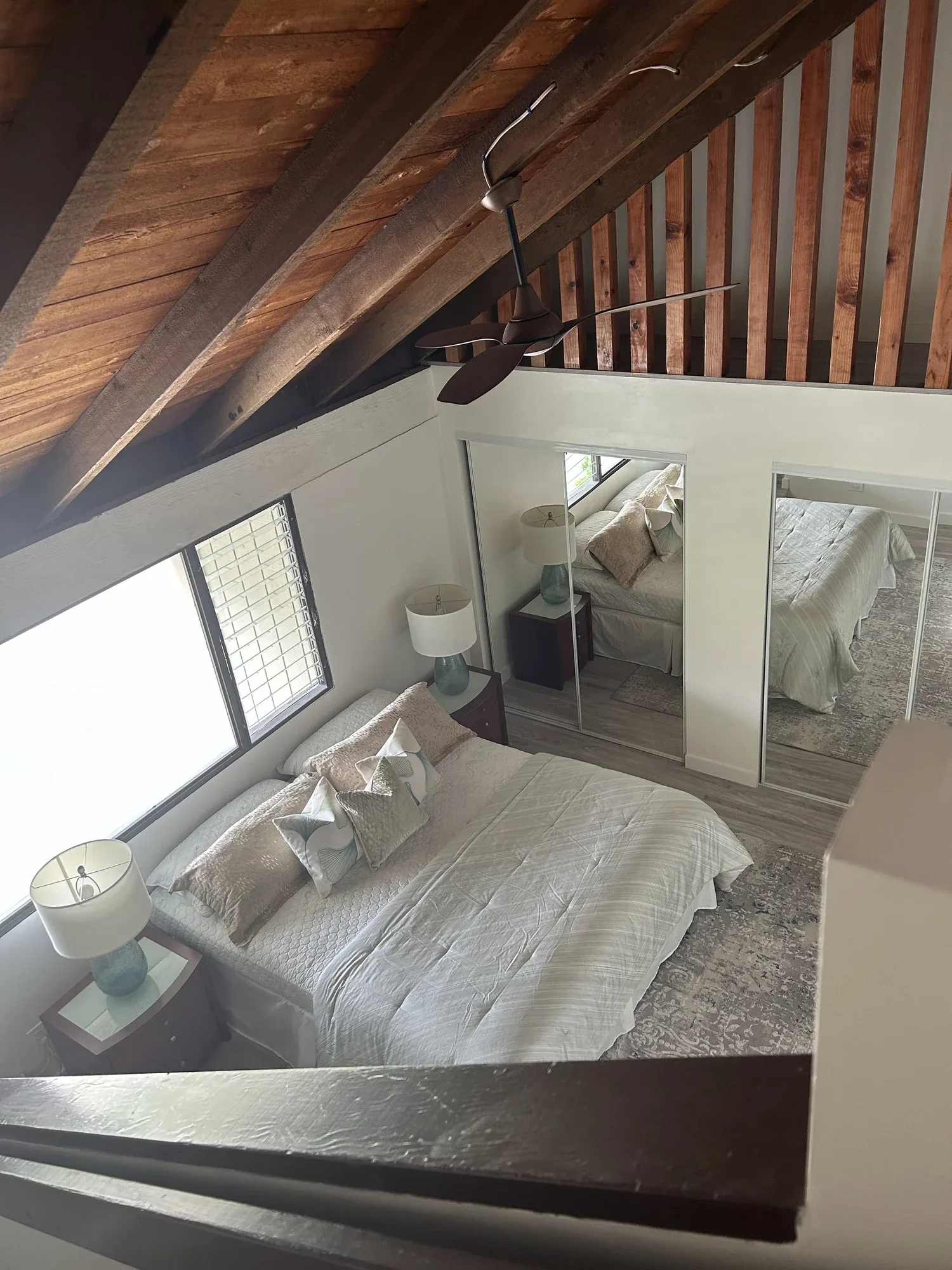 A bedroom with a sloped wooden ceiling, ceiling fan, large window, mirror closet doors, a bed with white bedding and pillows, and two nightstands with lamps.