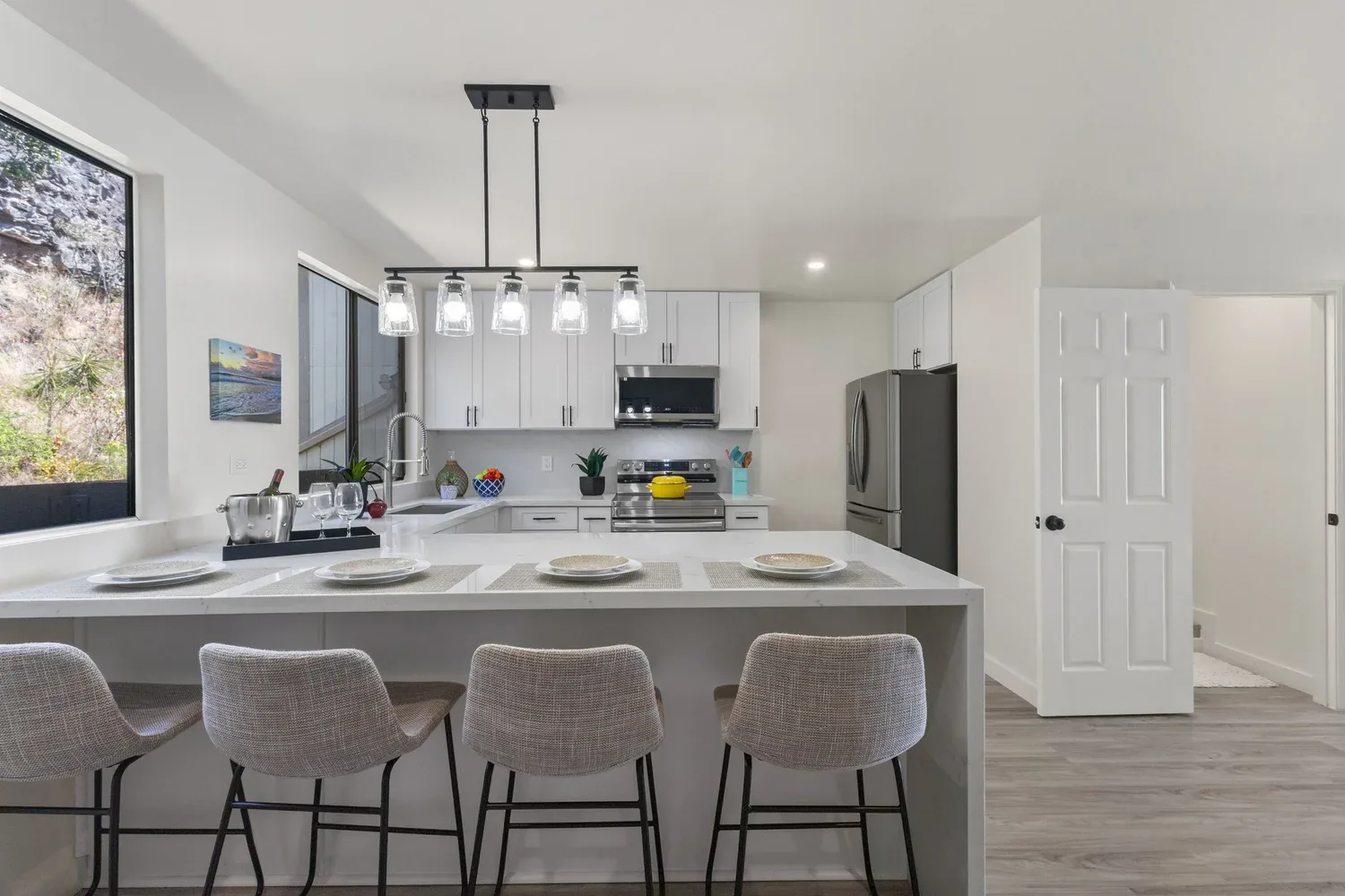 Modern kitchen with white cabinets, stainless steel appliances, an island with four beige chairs, and a pendant light fixture with six glass shades.