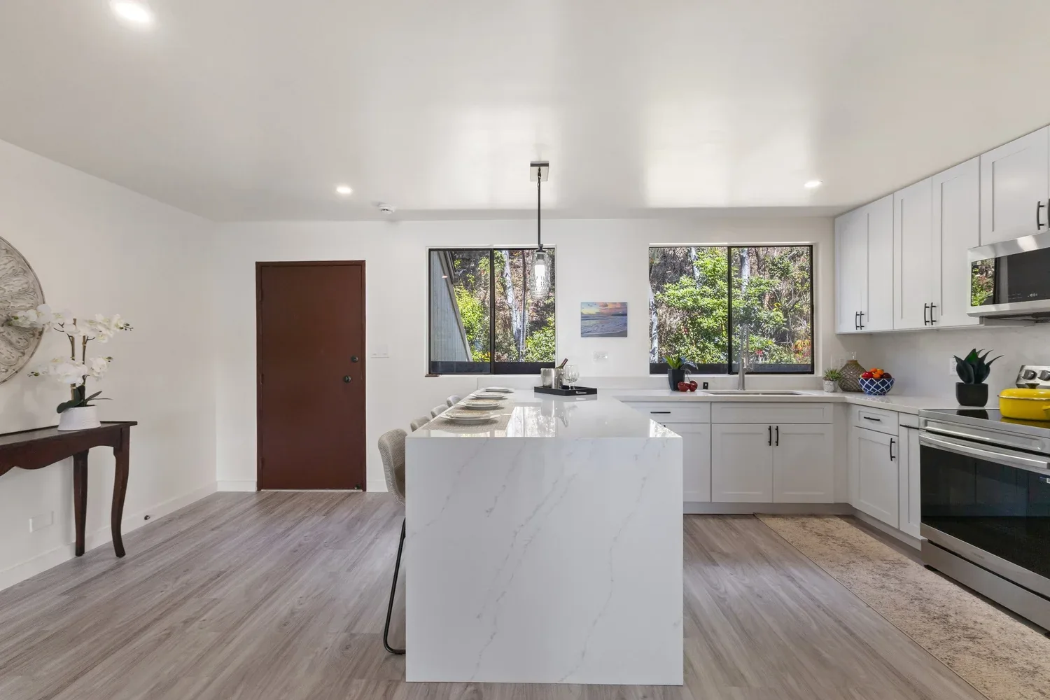 Modern kitchen with white cabinetry, marble island, and large windows overlooking greenery.